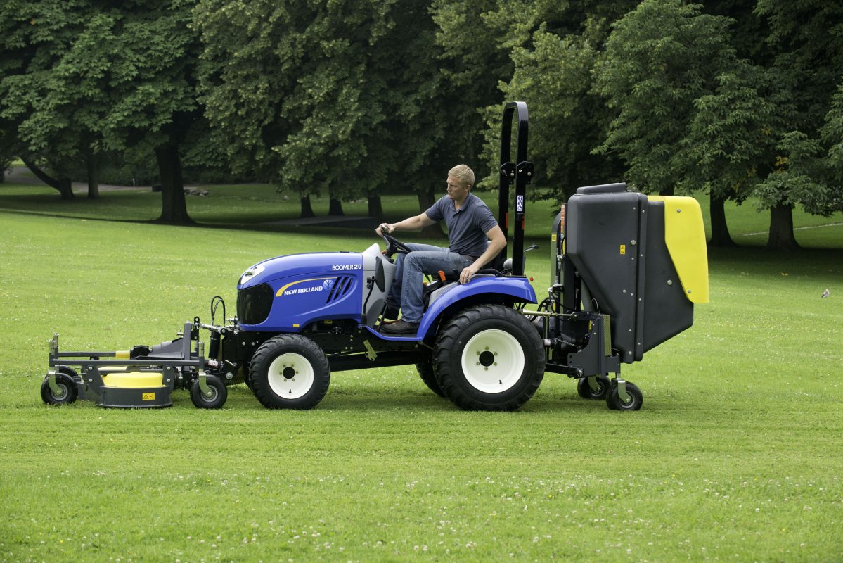 Ein blauer New Holland Boomer 20 Traktor mit Matev Frontmähwerk und gelbem Materialaufnahmegerät mäht eine große Rasenfläche in einem Park. Der Fahrer konzentriert sich auf die Arbeit, umgeben von dichtem Baumbestand und grüner Natur.