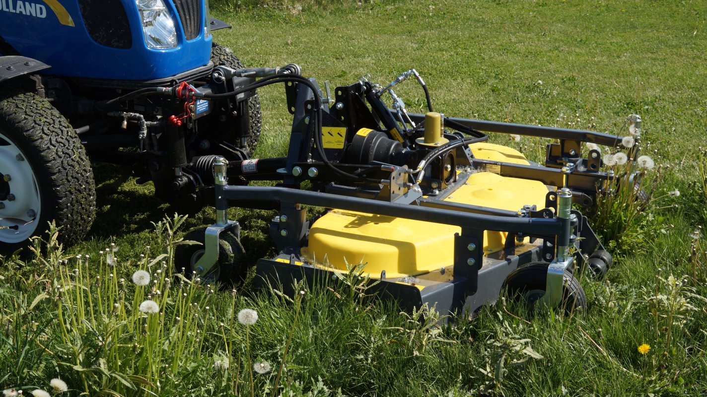 Nahaufnahme eines Matev Frontmähwerks mit gelber Verkleidung, montiert an einem blauen New Holland Traktor. Das Mähwerk befindet sich auf einer grünen Wiese mit Pusteblumen und zeigt hydraulische und mechanische Details.