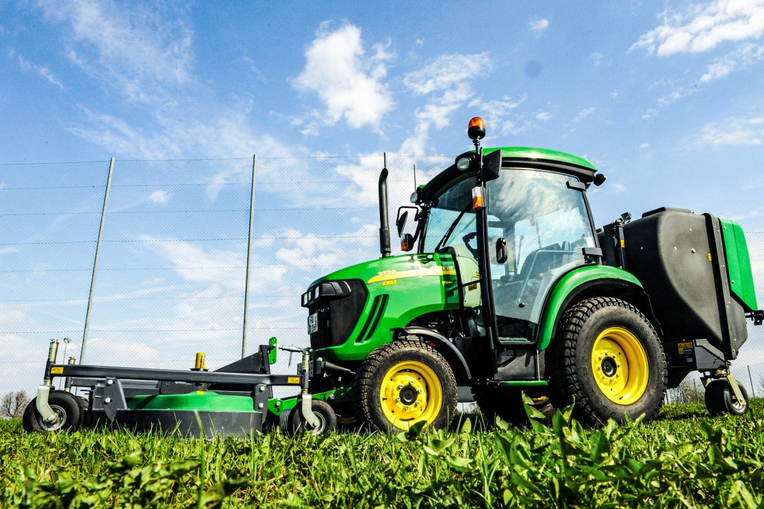 Grüner John Deere Kompakttraktor Modell 3720, ausgestattet mit einem matev Frontmähwerk und Grasaufnahmegerät, auf einer Wiese vor blauem Himmel mit vereinzelten Wolken.