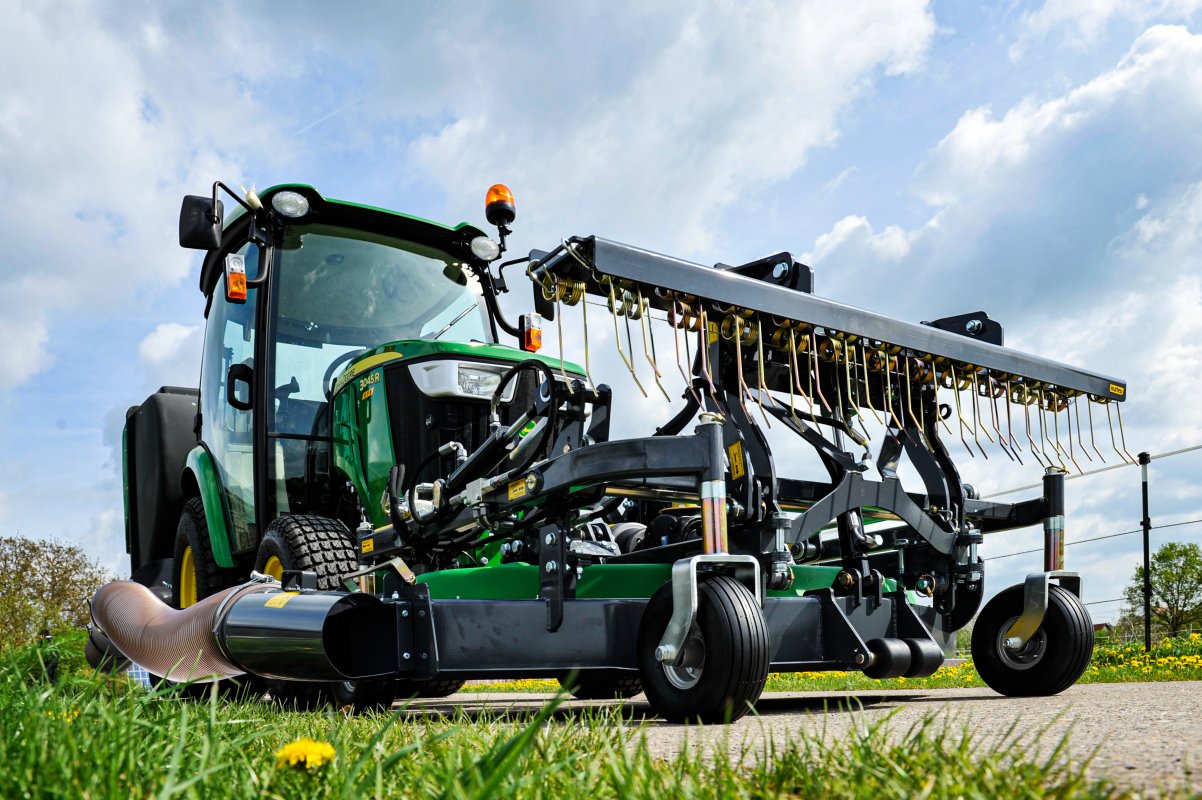 Ein John Deere 3046R Traktor mit Matev Frontmähwerk und Striegel, aufgenommen aus einer niedrigen Perspektive. Das Mähwerk zeigt detaillierte Striegelzinken und robuste Technik, während der Traktor auf einer Rasenfläche unter einem teilweise bewölkten Himmel steht.