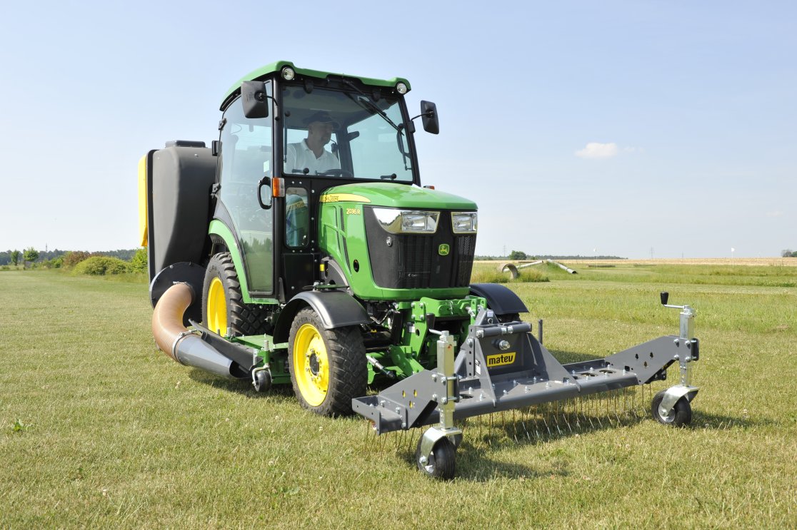 Ein John Deere 2036R Traktor mit einem Matev Rasenstriegel SCA 130 und Materialaufnahmegerät auf einer großen Rasenfläche. Der Traktor ist mit einem Schlauch verbunden, der das gesammelte Material in den Behälter leitet, während im Hintergrund eine weite, offene Landschaft sichtbar ist.
