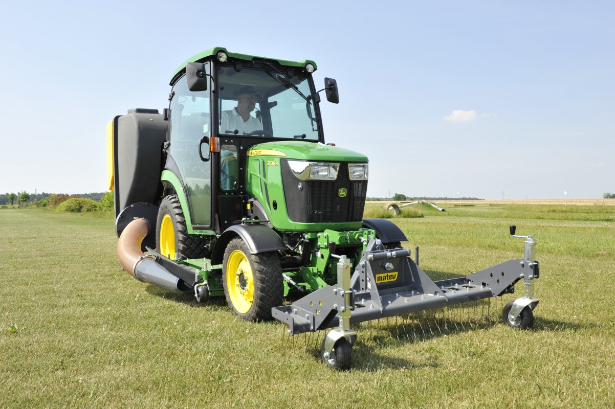 Ein John Deere 2036R Traktor mit einem Matev Rasenstriegel SCA 130 und Materialaufnahmegerät auf einer großen Rasenfläche. Der Traktor ist mit einem Schlauch verbunden, der das gesammelte Material in den Behälter leitet, während im Hintergrund eine weite, offene Landschaft sichtbar ist.