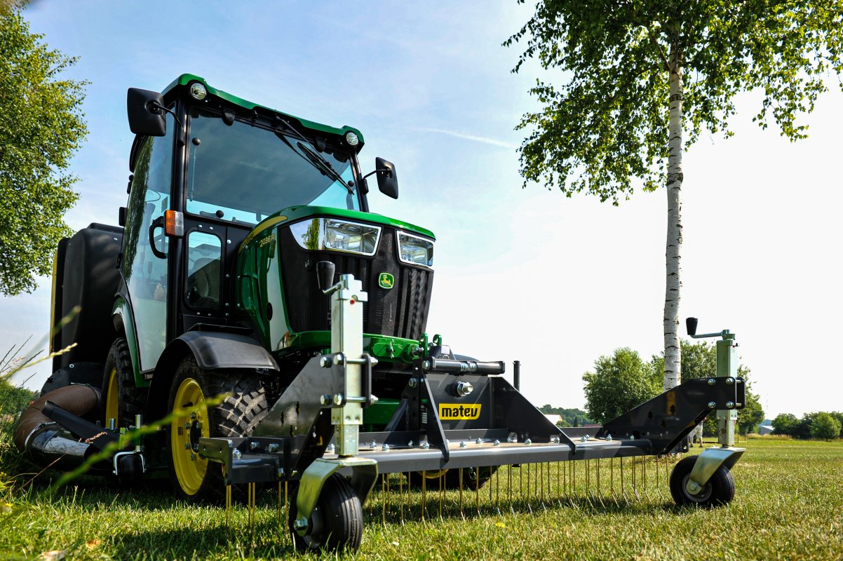 Ein John Deere 2036R Traktor mit einem Matev Rasenstriegel SCA 130 auf einer gepflegten Wiese. Der Striegel zeigt seine robusten Zinken, während im Hintergrund eine Birke und eine grüne Landschaft zu sehen sind, unter einem klaren blauen Himmel.