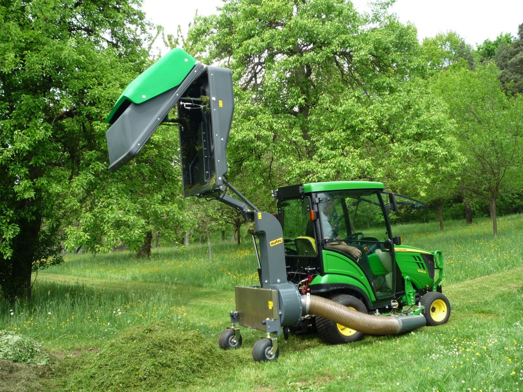 John Deere 1026R Traktor mit matev Grasaufnahmegerät, der geschnittenes Gras in einem Park oder Obstgarten entlädt. Effiziente Rasen- und Landschaftspflege inmitten grüner Natur mit Bäumen im Hintergrund.