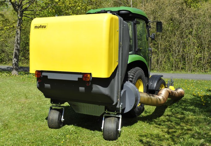 John Deere 3045 Traktor mit matev Grasaufnahmegerät in Gelb, ausgestattet mit Saugschlauch und Grasbehälter, auf einer grünen Wiese in ländlicher Umgebung. Effiziente Rasenpflege für professionelle Anwendungen.