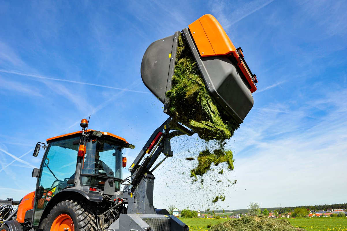 Kubota STW40 Traktor mit angebautem Grasaufnahmegerät von matev, das geschnittenes Gras auf ein Feld entlädt. Perfektes Zusammenspiel von Effizienz und Leistung bei der Rasenpflege vor einem klaren blauen Himmel und ländlicher Kulisse.