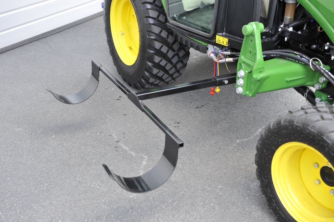 Ein Schlauchkellenhalter, montiert an einem John Deere 2026R Traktor. Die Halterung ist in schwarzer Farbe und an der Heckhydraulik des Traktors befestigt. Im Hintergrund ist ein Teil des Fahrzeugs mit gelben Rädern und schwarzen Reifen zu sehen. Der Traktor steht auf einem asphaltierten Untergrund.