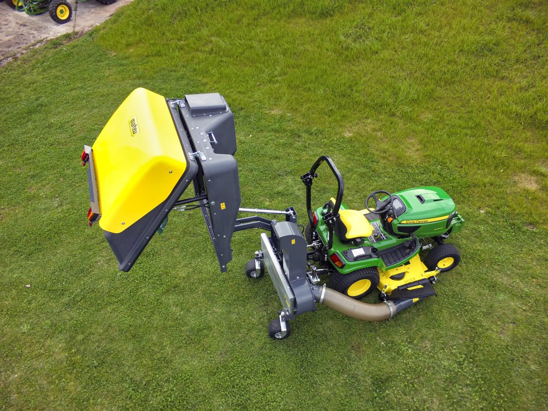 Ein John Deere X940 Aufsitzrasenmäher mit einem gelben Matev Materialaufnahmegerät in einer hochgekippten Position, aufgenommen von oben. Der Rasenmäher steht auf einer grünen Wiese, und im Hintergrund sind Teile weiterer Fahrzeuge sichtbar.