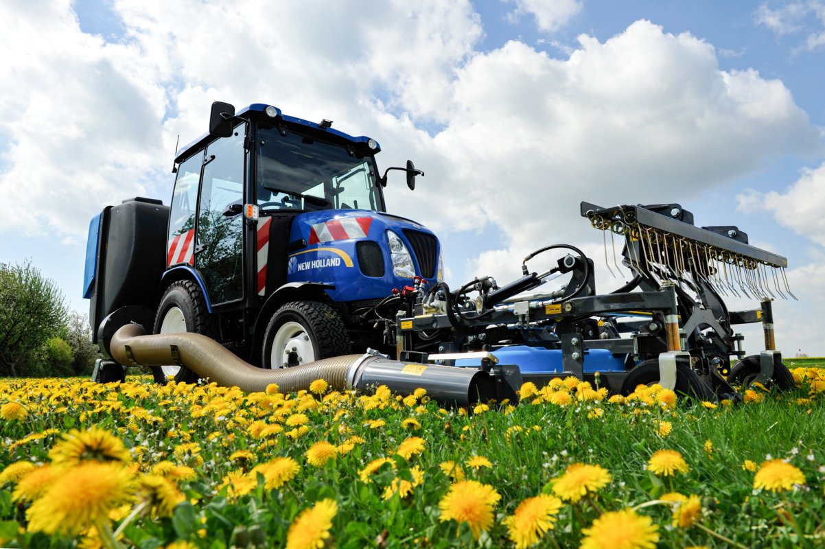 Ein blauer New Holland Traktor mit Matev Frontmähwerk und Striegel im Einsatz auf einer Wiese voller gelber Löwenzahnblüten. Die Aufnahme zeigt das Mähwerk aus nächster Nähe, während der Himmel teilweise bewölkt ist.