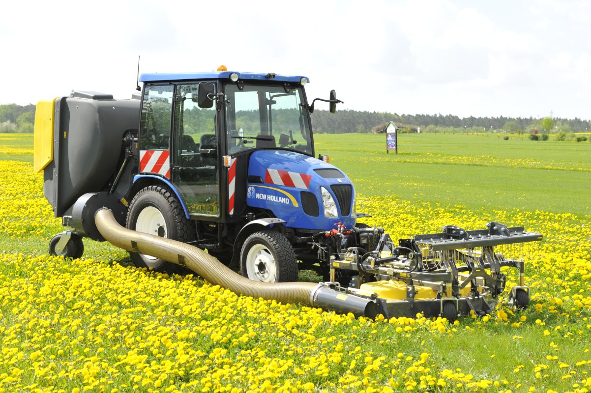 Ein blauer New Holland Traktor mit Matev Frontmähwerk und Materialaufnahmegerät mäht eine Wiese voller gelber Löwenzahnblüten. Im Hintergrund erstreckt sich eine grüne Landschaft mit vereinzelten Bäumen und einem bewölkten Himmel.