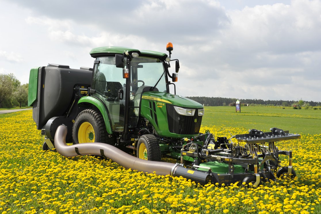Ein John Deere 3045R Traktor mit Matev Frontmähwerk und Materialaufnahmegerät mäht eine Wiese voller gelber Löwenzahnblüten. Die Szene zeigt einen weiten Hintergrund mit Feldern und Bäumen unter einem bewölkten Himmel.
