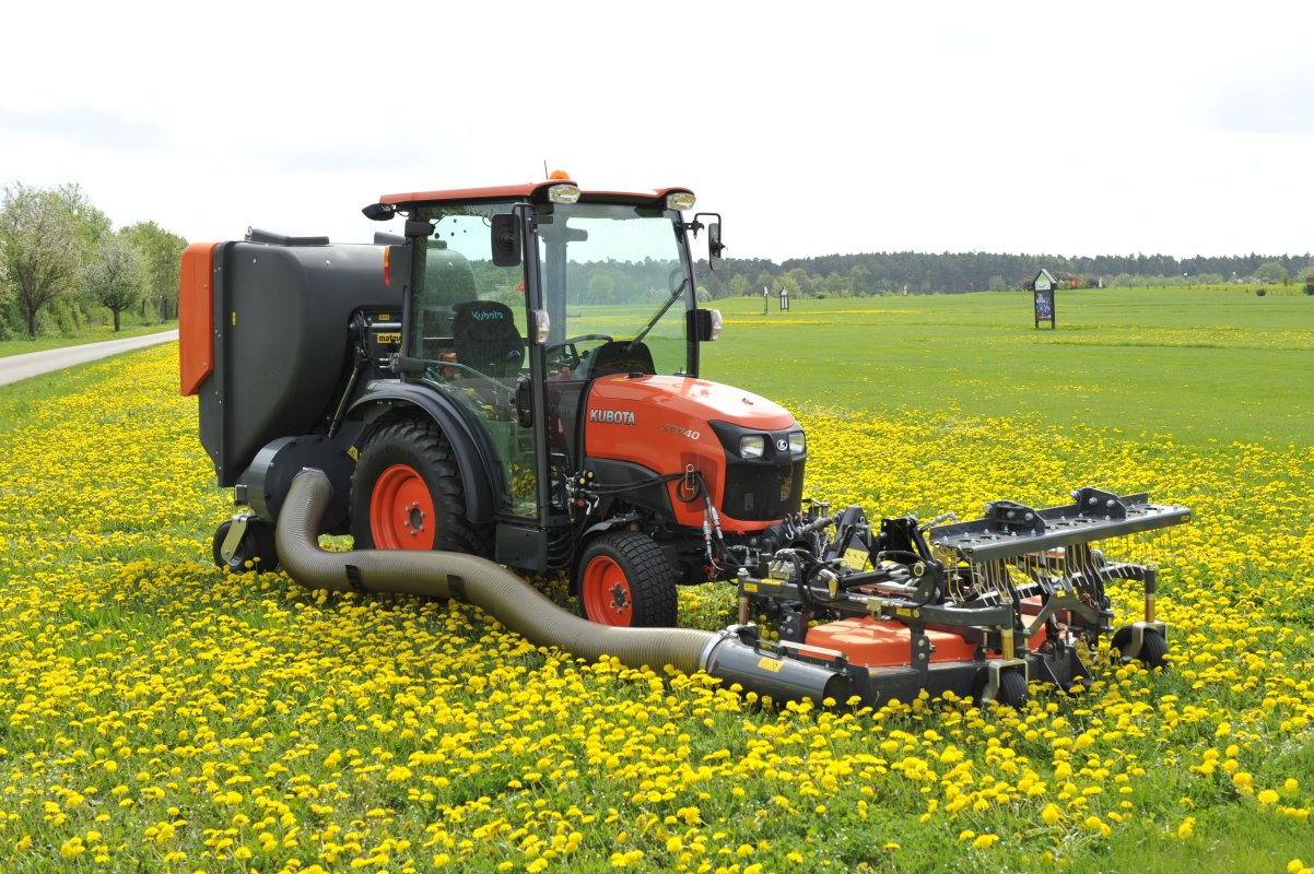 Ein Kubota STW40 Traktor mit Matev Frontmähwerk und Materialaufnahmegerät mäht eine Wiese voller blühender gelber Löwenzahnblüten. Im Hintergrund sind eine Straße, Bäume und ein offenes Feld zu sehen, unter einem bewölkten Himmel.