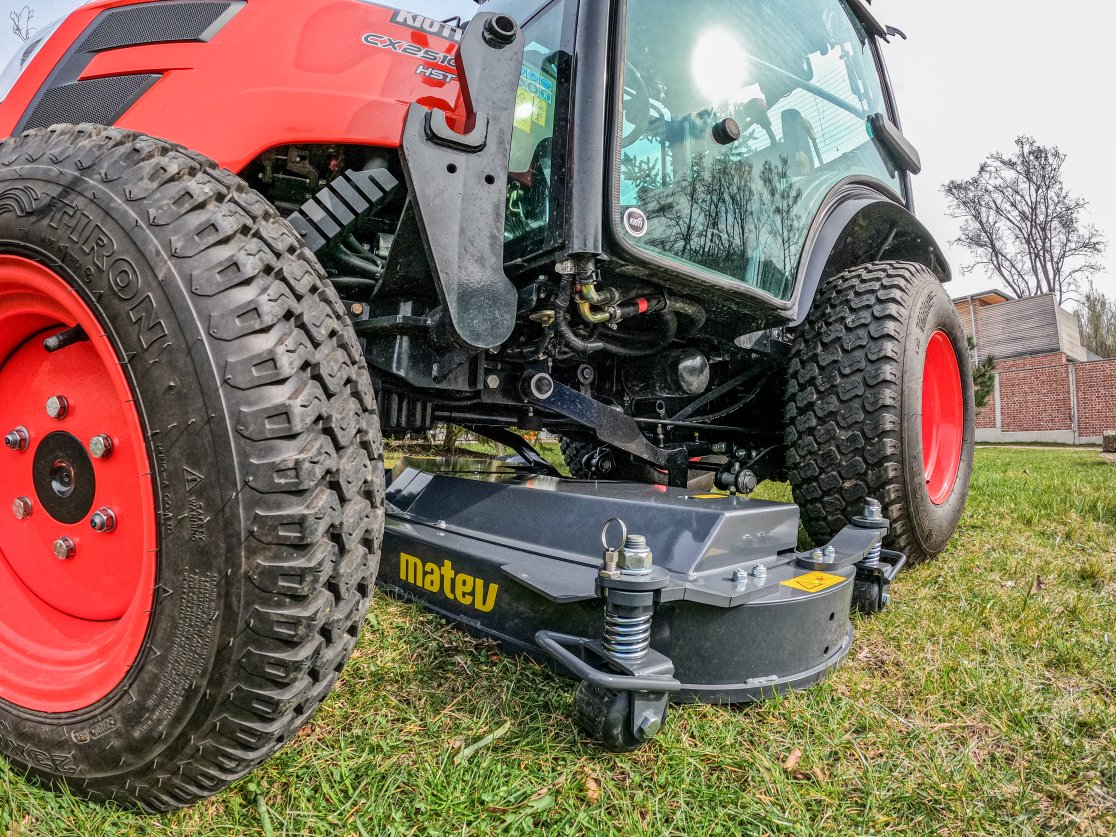Nahaufnahme eines matev MOW-MM 152 Multifunktionsmähwerks, angebracht an einem roten KIOTI CX2510 Traktor. Das Mähwerk und die großen Reifen des Traktors befinden sich auf einer grünen Wiese. Im Hintergrund sind Gebäude und Bäume sichtbar.