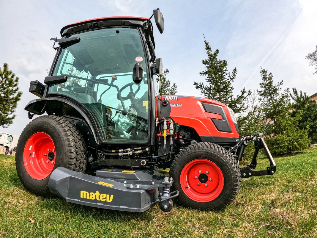 Ein Kioti CX2510 Traktor mit einem Matev Multifunktionsmähwerk auf einer grünen Wiese. Der Traktor ist seitlich abgebildet, mit einem klaren Blick auf die Kabine und die robuste Bauweise, umgeben von Bäumen und leicht bewölktem Himmel.