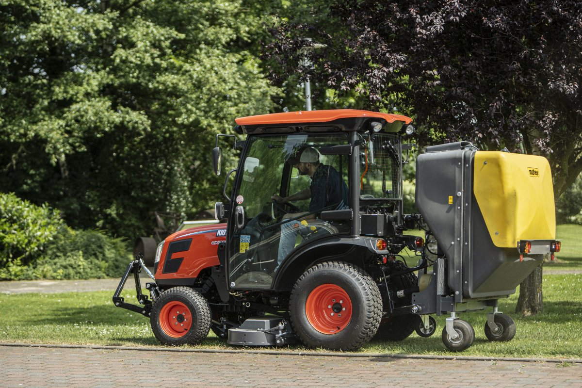 © Daedong Kioti Europe B.V. Ein Kioti CX2510 Traktor mit Matev Multifunktionsmähwerk und gelbem Materialaufnahmegerät im Einsatz auf einer gepflegten Rasenfläche. Der Fahrer arbeitet konzentriert, umgeben von dichter grüner Vegetation und einem Baum im Hintergrund. | © Daedong Kioti Europe B.V.