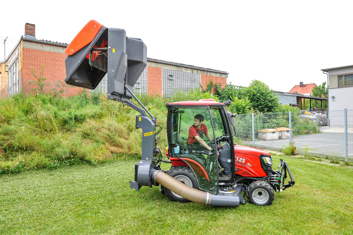 TYM TS25 Traktor mit matev Grasaufnahmegerät, der geschnittenes Gras in den Behälter lädt, während ein Fahrer in einer urbanen Umgebung mit grüner Wiese und Gebäuden im Hintergrund arbeitet. Effiziente Lösung für die professionelle Grundstückspflege.