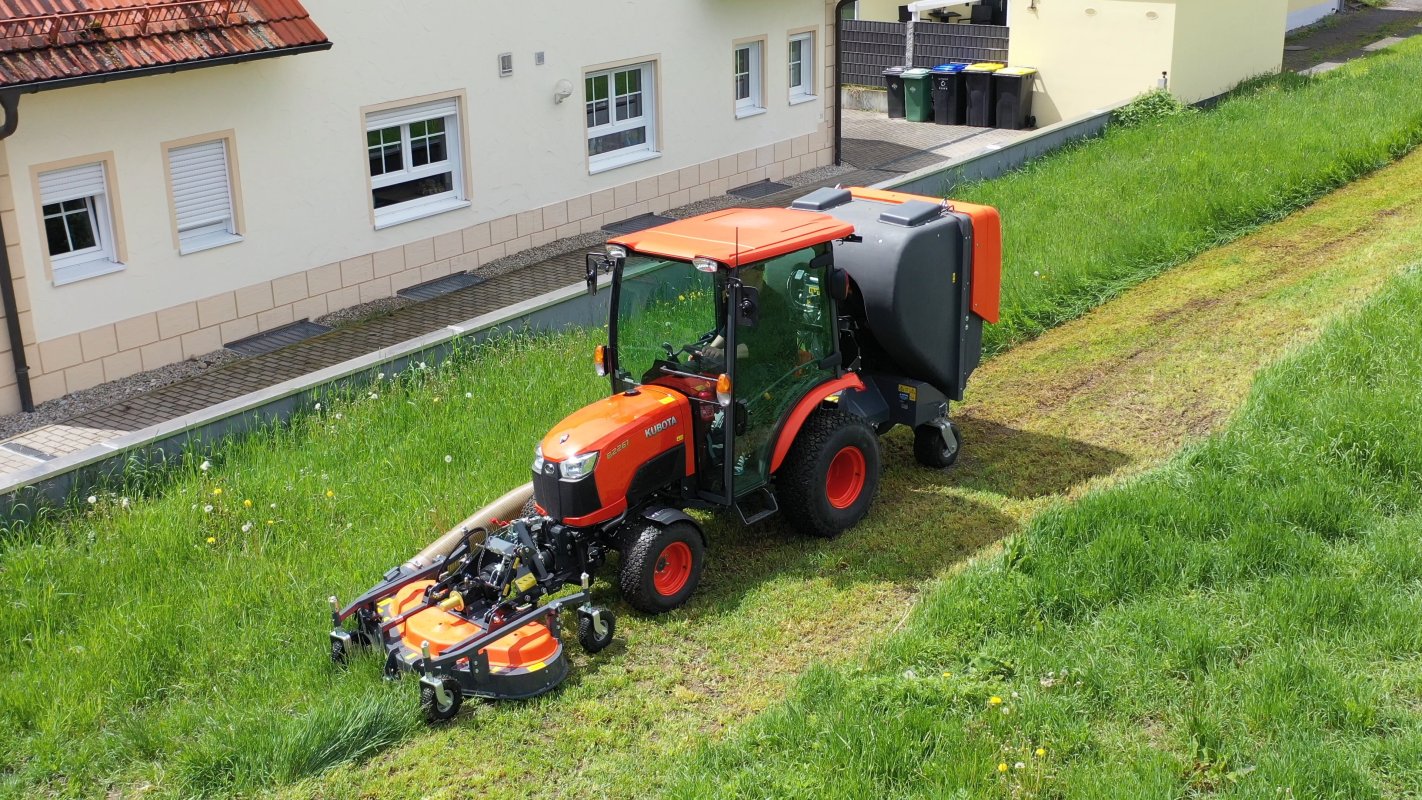 Ein Kubota B2261 Traktor mit Matev Frontmähwerk und Grasaufnahmegerät im Einsatz auf einer Wiese vor einem Wohnhaus. Das gemähte Gras wird effizient aufgenommen, während die Umgebung durch saftiges Grün und gepflegte Wege geprägt ist.