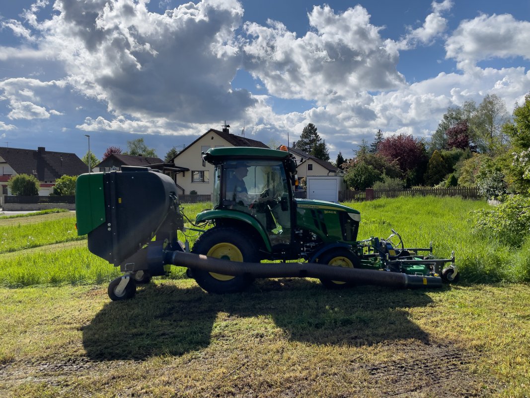 Ein John Deere 3045R Traktor mit einem Matev Frontmähwerk und Materialaufnahmegerät im Einsatz auf einer Wiese. Im Hintergrund befinden sich Wohnhäuser und Bäume, während der Himmel mit eindrucksvollen Wolkenformationen bedeckt ist.