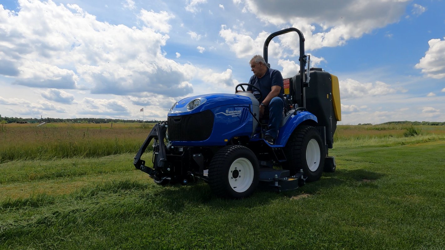 Ein blauer New Holland Boomer 25 Traktor mit einem Matev Zwischenachsmähwerk und Materialaufnahmegerät im Einsatz auf einer grünen Wiese. Der Fahrer sitzt konzentriert am Steuer, während der Himmel von weißen Wolken durchzogen ist.