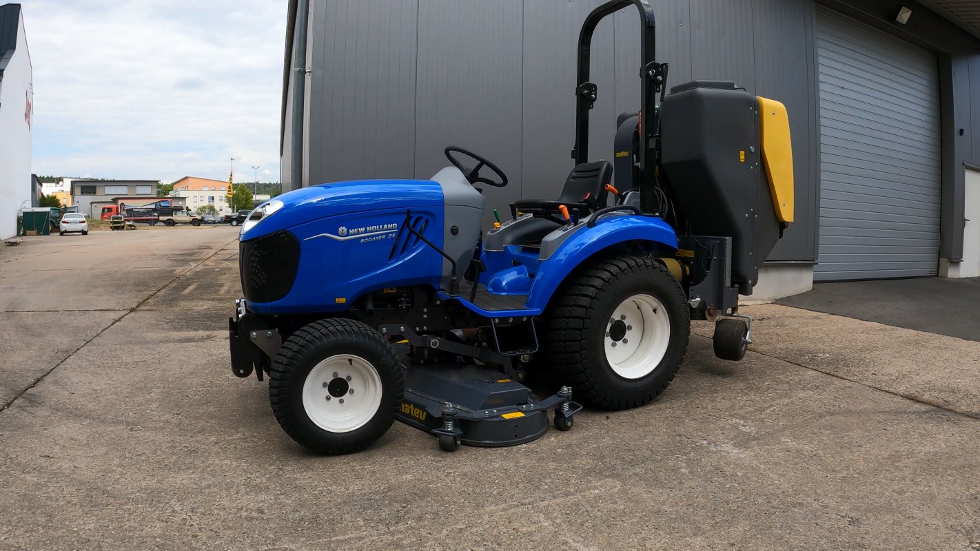 Ein blauer New Holland Boomer 25 Traktor mit einem Matev Zwischenachsmähwerk und Materialaufnahmegerät steht auf einem betonierten Platz vor einem Industriegebäude. Im Hintergrund sind weitere Fahrzeuge und Gebäude zu sehen.