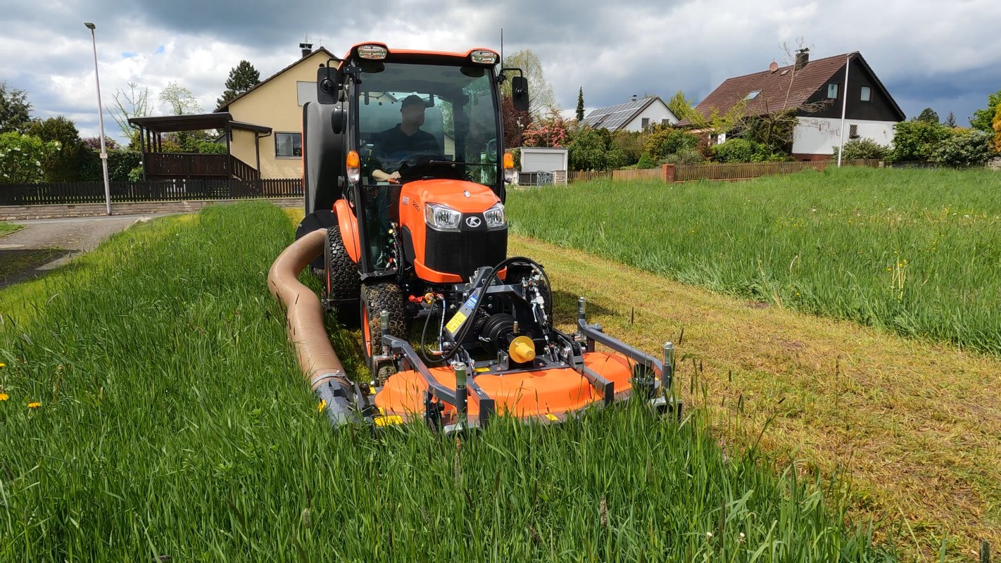 Ein Kubota Traktor mit einem Matev Frontmähwerk und einem Grasaufnahmegerät im Einsatz auf einer Wiese, umgeben von Wohnhäusern und dichter Vegetation unter einem teils bewölkten Himmel.