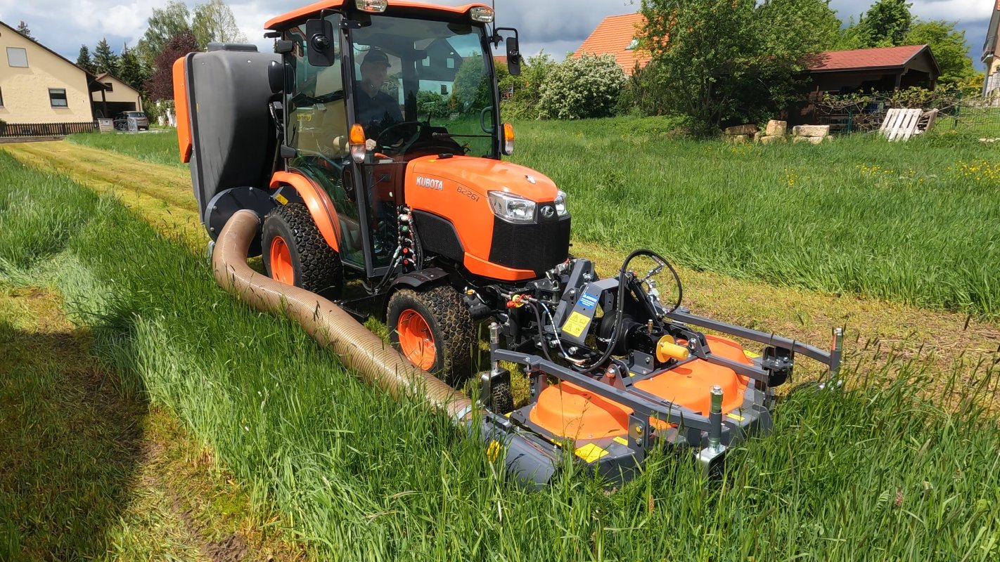 Ein Kubota B2261 Traktor mit einem Matev Frontmähwerk und Grasaufnahmegerät mäht hohes Gras auf einer Wiese. Im Hintergrund sind Wohnhäuser und eine dichte Vegetation sichtbar, während der Schlauch für die Grasaufnahme das gemähte Gras effizient transportiert.