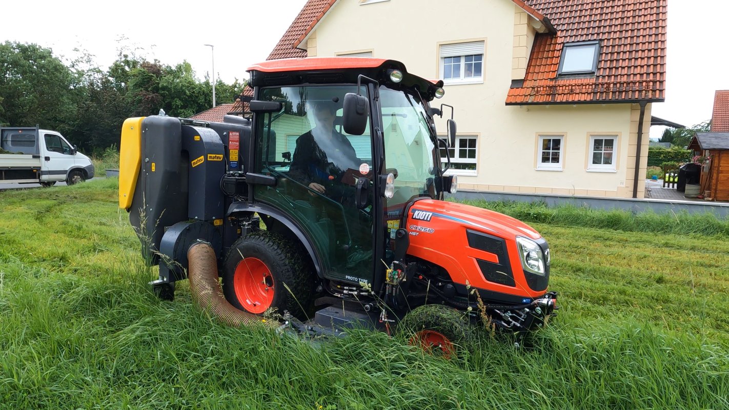 Ein Kioti CX2510 HST Traktor mit einem Matev Zwischenachsmähwerk und Materialaufnahmegerät mäht hohes Gras vor einem Wohnhaus mit rotem Ziegeldach. Im Hintergrund sind ein Transporter und eine grüne Gartenlandschaft zu sehen.