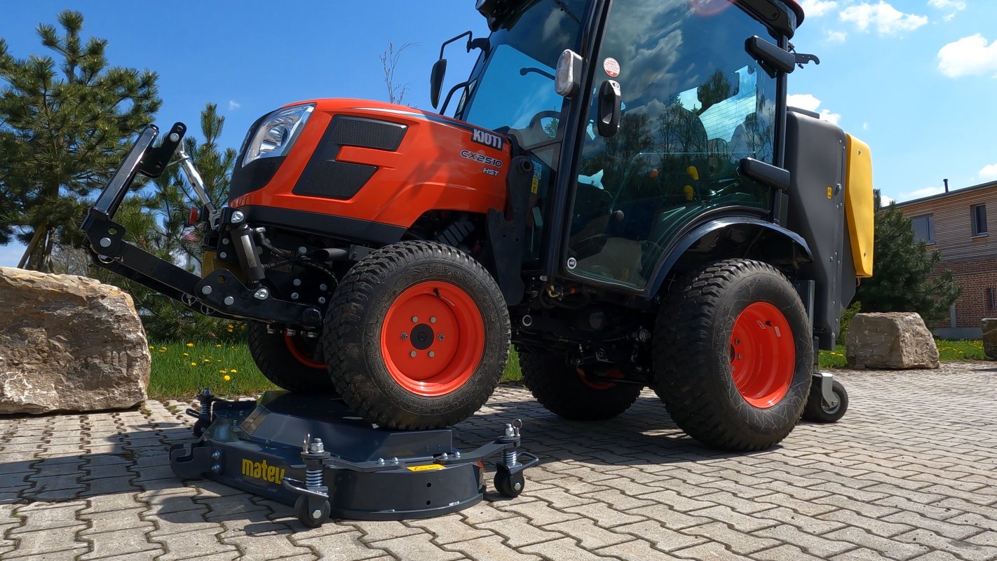 Ein Kioti CX2510 HST Traktor mit einem Matev Zwischenachsmähwerk und Materialaufnahmegerät, geparkt auf einem gepflasterten Untergrund. Im Hintergrund sind Bäume, ein Gebäude und Felsbrocken zu sehen, die eine natürliche Umgebung schaffen.