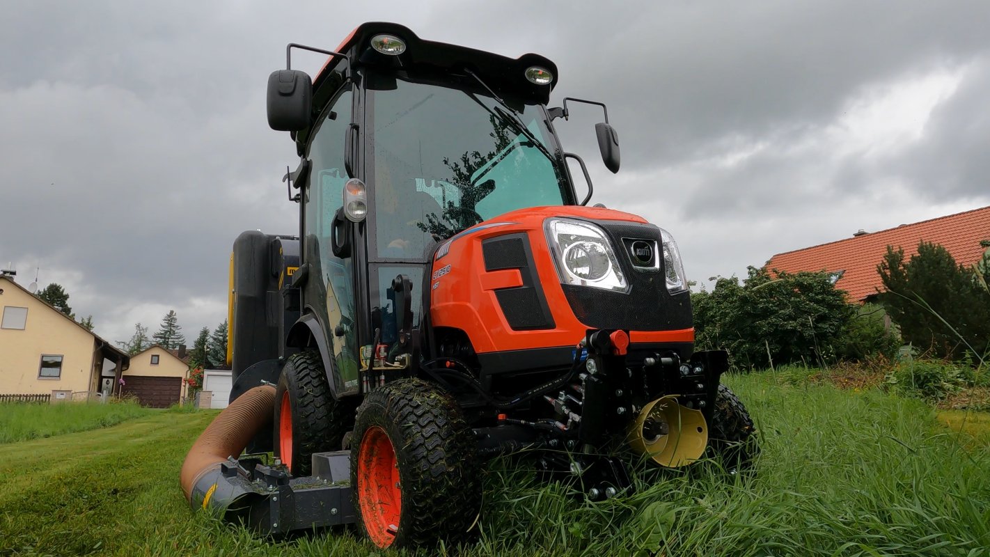 Ein Kioti CX2510 HST Traktor mit einem Matev Zwischenachsmähwerk und Materialaufnahmegerät auf einer Wiese vor einem Wohngebiet. Der Himmel ist bedeckt mit dunklen Wolken, was auf regnerisches Wetter hindeutet.