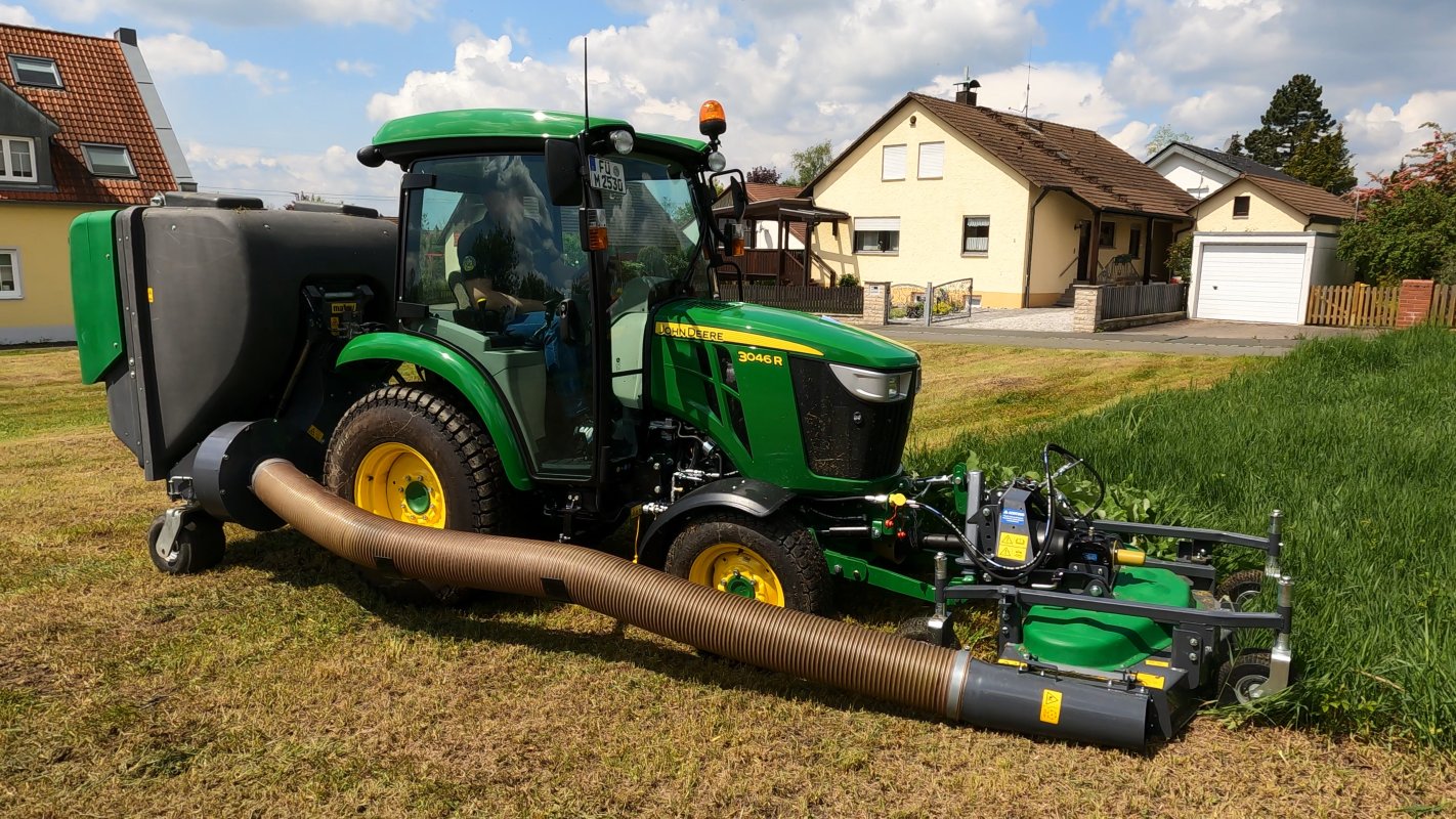 Ein John Deere 3046R Traktor mit einem Matev Frontmähwerk und Materialaufnahmegerät im Einsatz auf einer Wiese vor Einfamilienhäusern. Der Traktor mäht das hohe Gras, während der Schlauch das gemähte Material in den Sammelbehälter leitet.