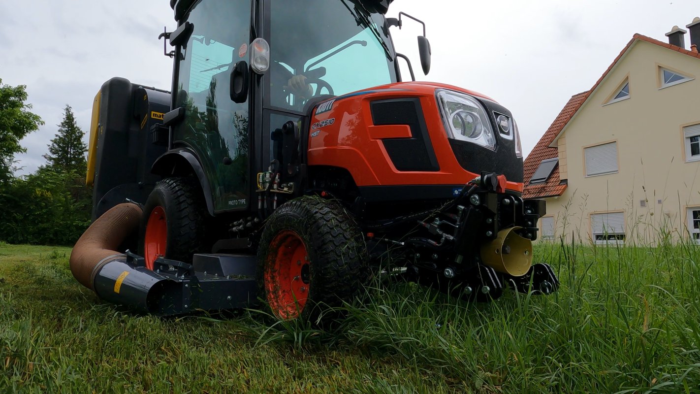 Ein Kioti CX2510 HST Traktor mit einem Matev Zwischenachsmähwerk und Materialaufnahmegerät mäht hohes Gras auf einer Wiese vor einem Wohnhaus mit roten Dachziegeln. Der Himmel ist bedeckt, und die Umgebung zeigt eine grüne, gepflegte Landschaft.