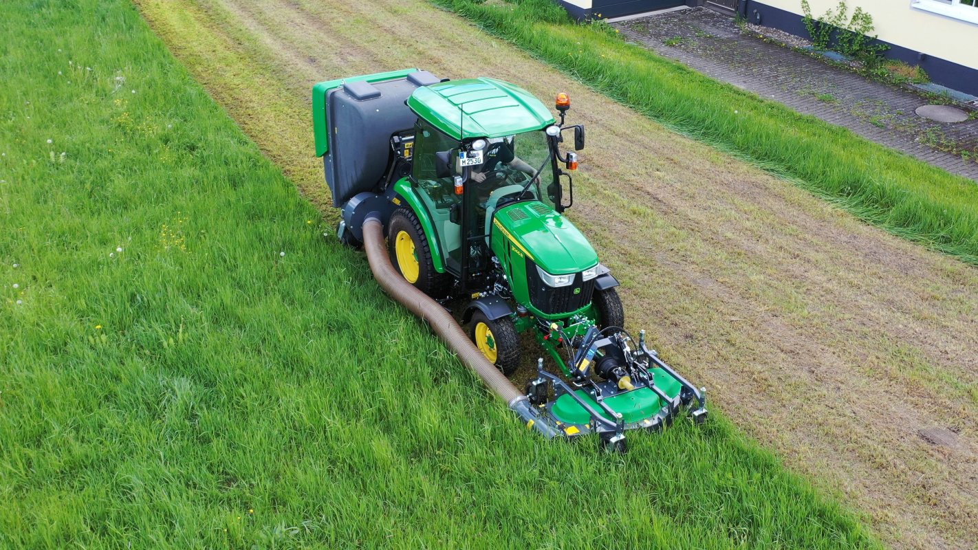 Ein John Deere 3045R Traktor mit einem Matev Frontmähwerk und Materialaufnahmegerät im Einsatz auf einer Wiese. Der Schlauch transportiert das gemähte Gras, während die Fläche präzise gemäht wird. Im Hintergrund ist eine gepflasterte Einfahrt sichtbar.