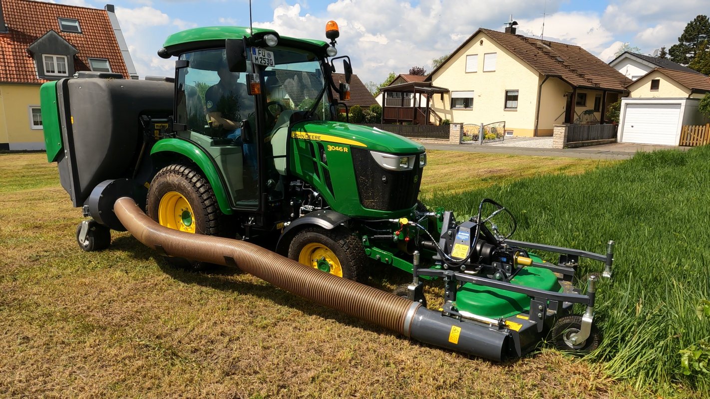 Ein John Deere 3045R Traktor mit einem Matev Frontmähwerk und Materialaufnahmegerät mäht hohes Gras vor einem Wohngebiet mit Einfamilienhäusern. Der Schlauch leitet das gemähte Gras in den Sammelbehälter, während der Traktor auf einer frisch gemähten Fläche steht.