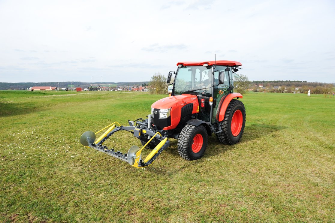 Ein roter Kubota L2-622 Traktor mit einem matev MOW-PT Portalmähwerk steht auf einer grünen Wiese. Im Hintergrund ist eine ländliche Umgebung mit Gebäuden, Bäumen und Windrädern zu sehen.