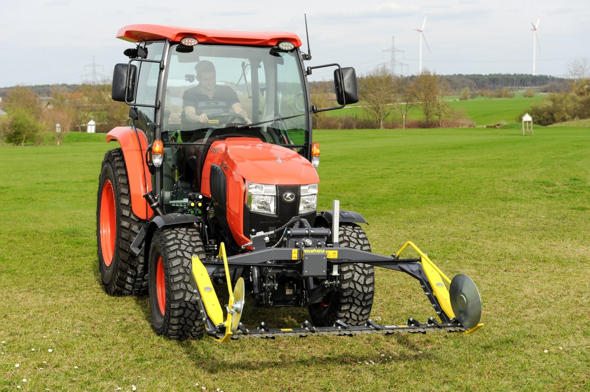 Ein Kubota L2-622 Traktor mit einem Matev Portalmähwerk, das auf einer großen grünen Wiese betrieben wird. Im Hintergrund sind Windräder und eine ländliche Landschaft unter einem leicht bewölkten Himmel zu sehen.
