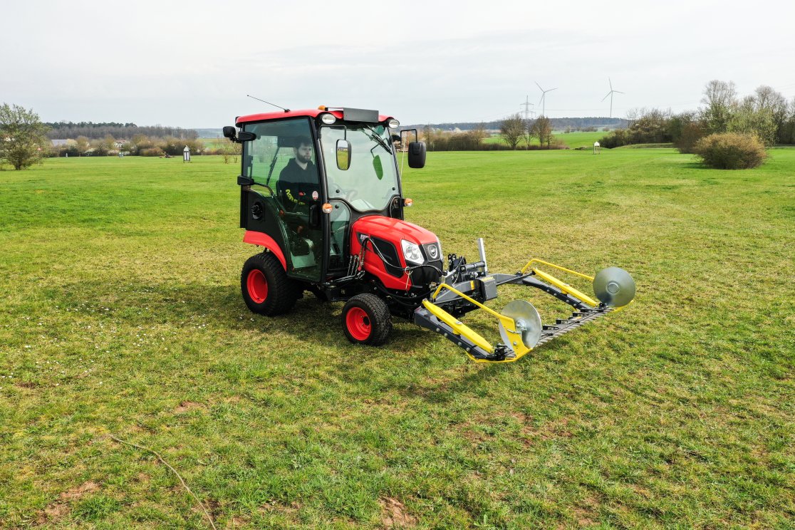 Ein roter Kioti CS2520 Traktor mit Kabine und einem angebauten matev MOW-PT Portalmähwerk steht auf einer weitläufigen Wiese. Im Hintergrund sind Windräder und eine ländliche Landschaft zu erkennen, die das Einsatzgebiet unterstreichen.