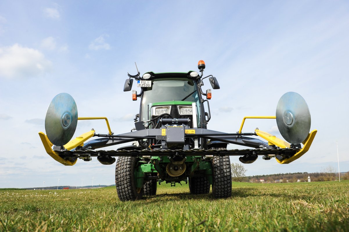 Ein John Deere Traktor der 3R-Serie mit einem Matev Portalmähwerk im Einsatz auf einer Wiese. Das Portalmähwerk ist frontal abgebildet, mit seinen großen gelben Schutzscheiben und klar sichtbarer Technik, unter einem leicht bewölkten Himmel.