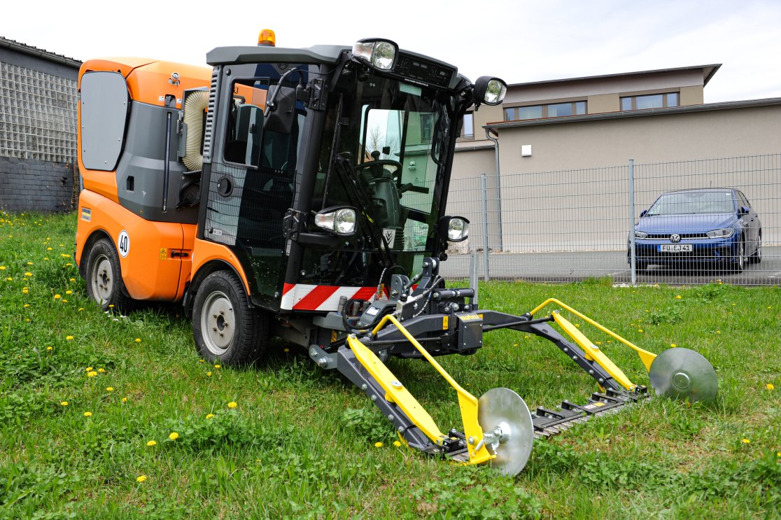 Eine orangefarbene Kommunalmaschine Kärcher MC 130 mit einem angebauten matev MOW-PT Portalmähwerk steht auf einer grünen Wiese mit blühendem Löwenzahn. Im Hintergrund sind ein Zaun, ein Gebäude und ein geparktes Auto sichtbar.