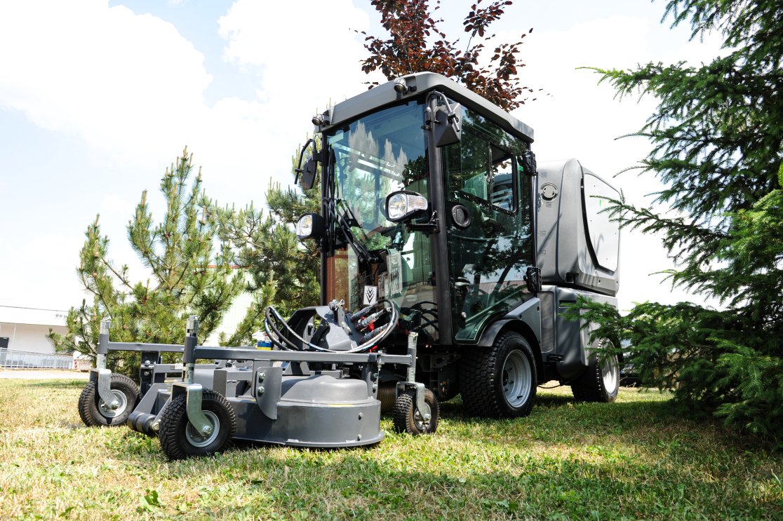 Eine Kommunalmaschine mit Kabine, ausgestattet mit einem matev MOW-FM 14 Frontmähwerk, steht auf einer gepflegten Rasenfläche. Im Hintergrund sind Nadelbäume und ein Industriegebäude unter einem blauen Himmel zu sehen.