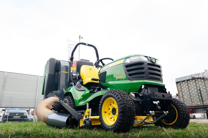 Ein John Deere X948 Aufsitzmäher mit matev-Anbaugerät, aufgenommen aus einer niedrigen Perspektive. Der Traktor ist mit einem Saugsystem ausgestattet, ideal für die Grünflächenpflege. Im Hintergrund sind eine Metallabsperrung und geparkte Fahrzeuge zu sehen.