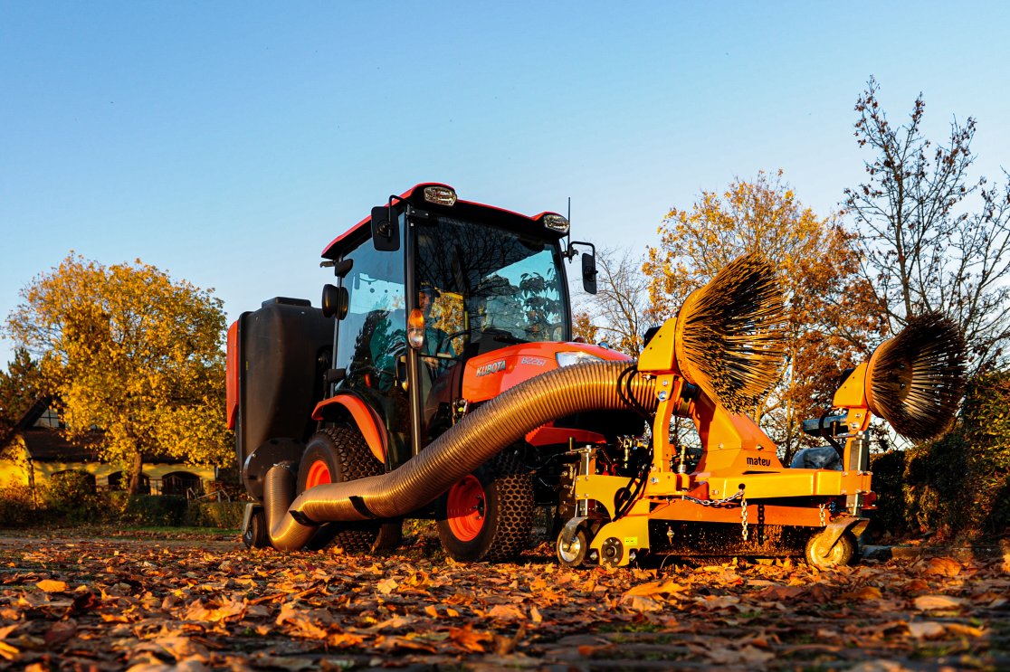 Ein Kubota B2261 Traktor in leuchtendem Orange mit einer angebauten matev Multifunktions-Saugdüse und Seitenbesen. Der Traktor arbeitet auf einem herbstlich bedeckten Gelände, umgeben von Bäumen in goldenen Herbstfarben. Der lange Saugschlauch und die rotierenden Bürsten heben sich hervor, während die Abendsonne die Szene erhellt.