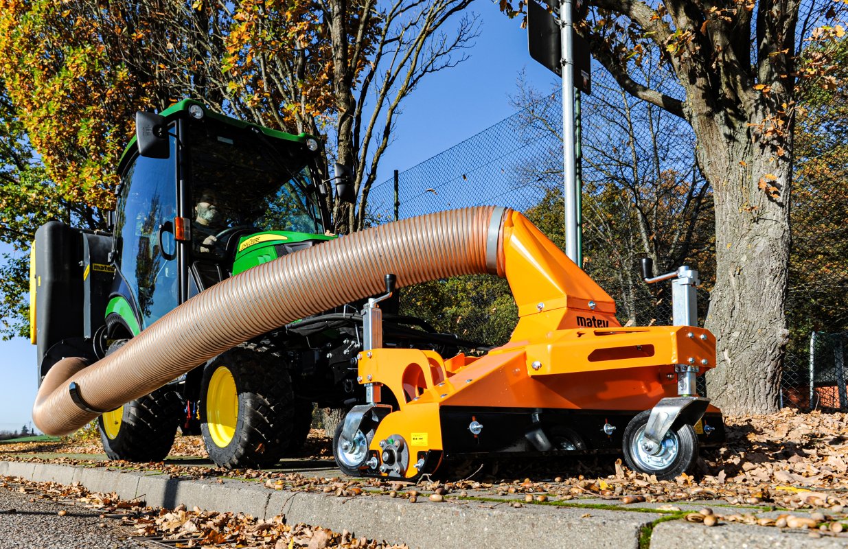 Ein kompaktes Multifunktions-Sauggerät, verbunden mit einem grünen John Deere Kompakttraktor. Das orangefarbene Gerät verfügt über einen flexiblen Saugschlauch und robuste Räder, ideal für die effiziente Aufnahme von Laub und Schmutz in kommunalen oder gartenbaulichen Bereichen.