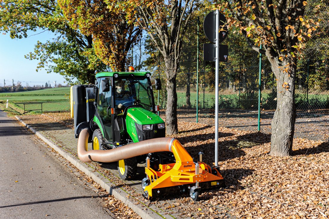 Ein grüner John Deere 2026R Traktor ist mit einer orangefarbenen matev-Multifunktionssaugdüse ausgestattet und auf einem mit Herbstlaub bedeckten Gehweg geparkt. Im Hintergrund stehen Bäume mit herbstlich gefärbtem Laub, und es ist ein Sportplatz mit einem Zaun sichtbar.