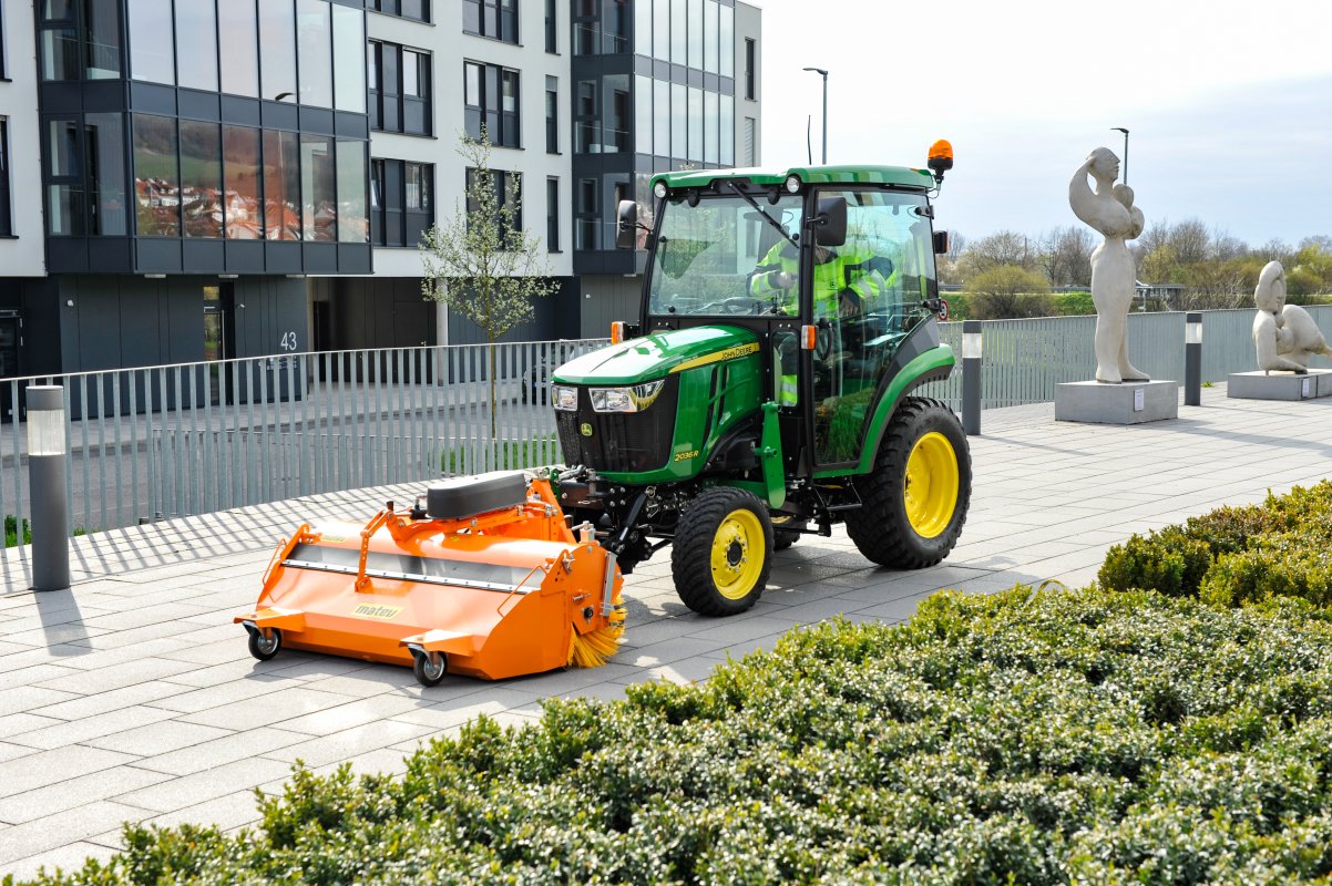 ohn Deere 2026R Traktor mit matev Kehrmaschine in Orange, reinigt moderne Pflasterflächen vor einem Bürogebäude mit Skulpturen und urbanem Umfeld. Perfekte Lösung für professionelle Flächenreinigung in städtischer Umgebung.