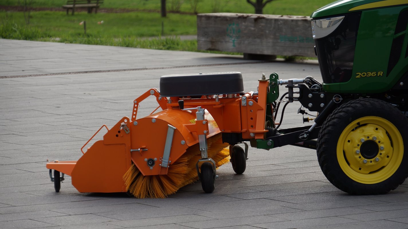 John Deere 2036R Traktor mit matev Kehrmaschine in Orange, ideal für die Reinigung von gepflasterten Flächen. Nahaufnahme der robusten Bürste und des Anbausystems, perfekt für professionelle Außenreinigung.