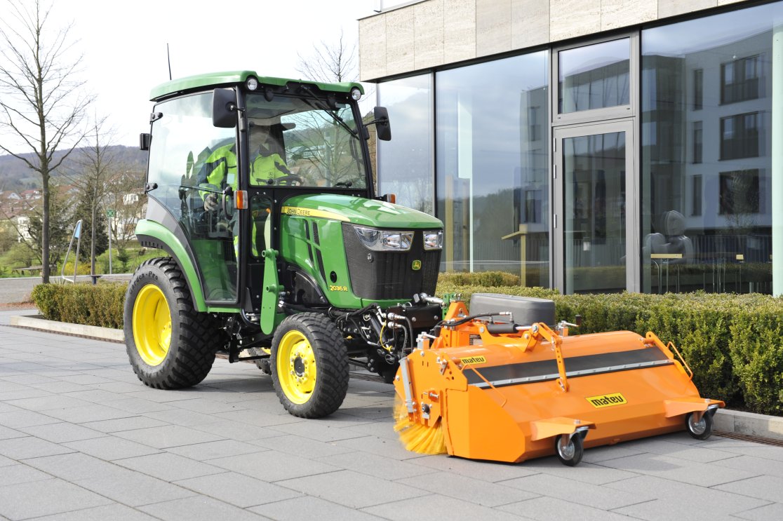 Ein grüner John Deere 2026R Traktor mit einer angehängten orangefarbenen matev-Kehrmaschine steht auf einer gepflasterten Fläche vor einem modernen Gebäude mit Glasfassade. Im Hintergrund sind gepflegte Hecken und eine leicht bewölkte Landschaft zu sehen.