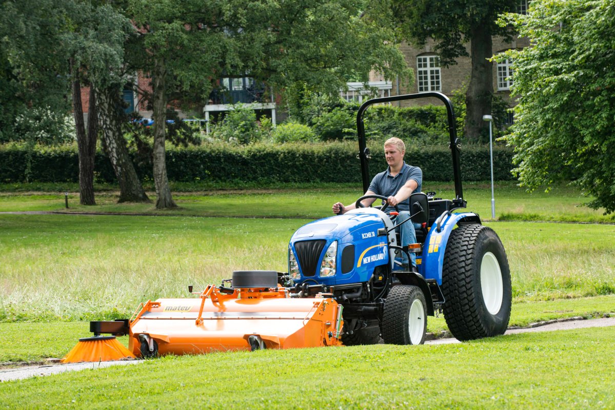 New Holland BO30 Traktor mit matev Kehrmaschine in Orange, bei der Pflege von Grünflächen in einem Park. Fahrer reinigt einen Weg, umgeben von Bäumen und Gebäuden im Hintergrund. Effiziente Lösung für professionelle Landschaftspflege.