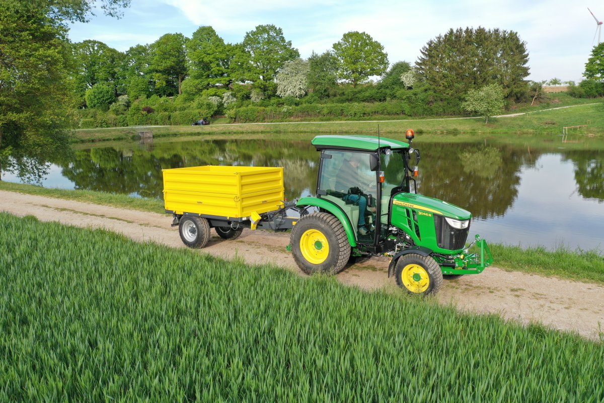 Ein grüner John Deere 3046R Traktor mit einem angehängten gelben Einachsdreiseitenkipper fährt auf einem Feldweg neben einem Teich. Die Szene ist von grüner Vegetation und einem klaren blauen Himmel umgeben. Im Hintergrund sind Bäume und eine Windkraftanlage zu sehen.