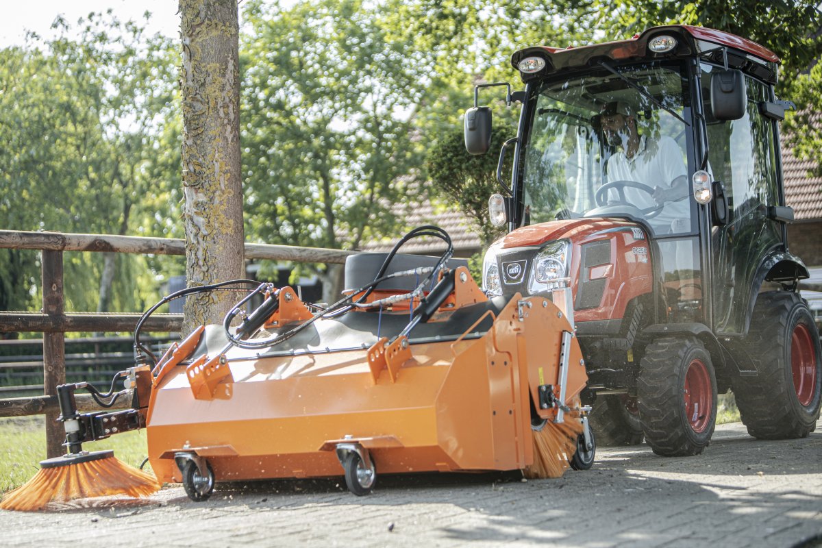 Kioti CX2510 Traktor mit matev Kehrmaschine in Orange, bei der Reinigung von Pflasterflächen neben einem Baum und einem Holzzaun. Ideal für professionelle Reinigungsarbeiten in ländlicher oder stadtnaher Umgebung.