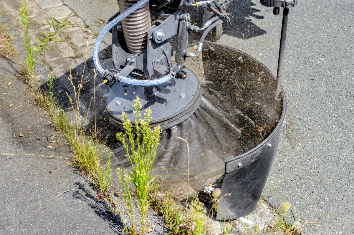 Detailaufnahme einer rotierenden Wildkrautbürste von matev im Einsatz entlang einer Bordsteinkante. Die Bürste wirbelt Schmutz und Unkraut auf, während Wasser zur Staubbindung eingesetzt wird. Im Vordergrund sind Pflanzen und Gräser zu sehen, die von der Bürste entfernt werden. Die Straße besteht aus Asphalt und Pflastersteinen.
